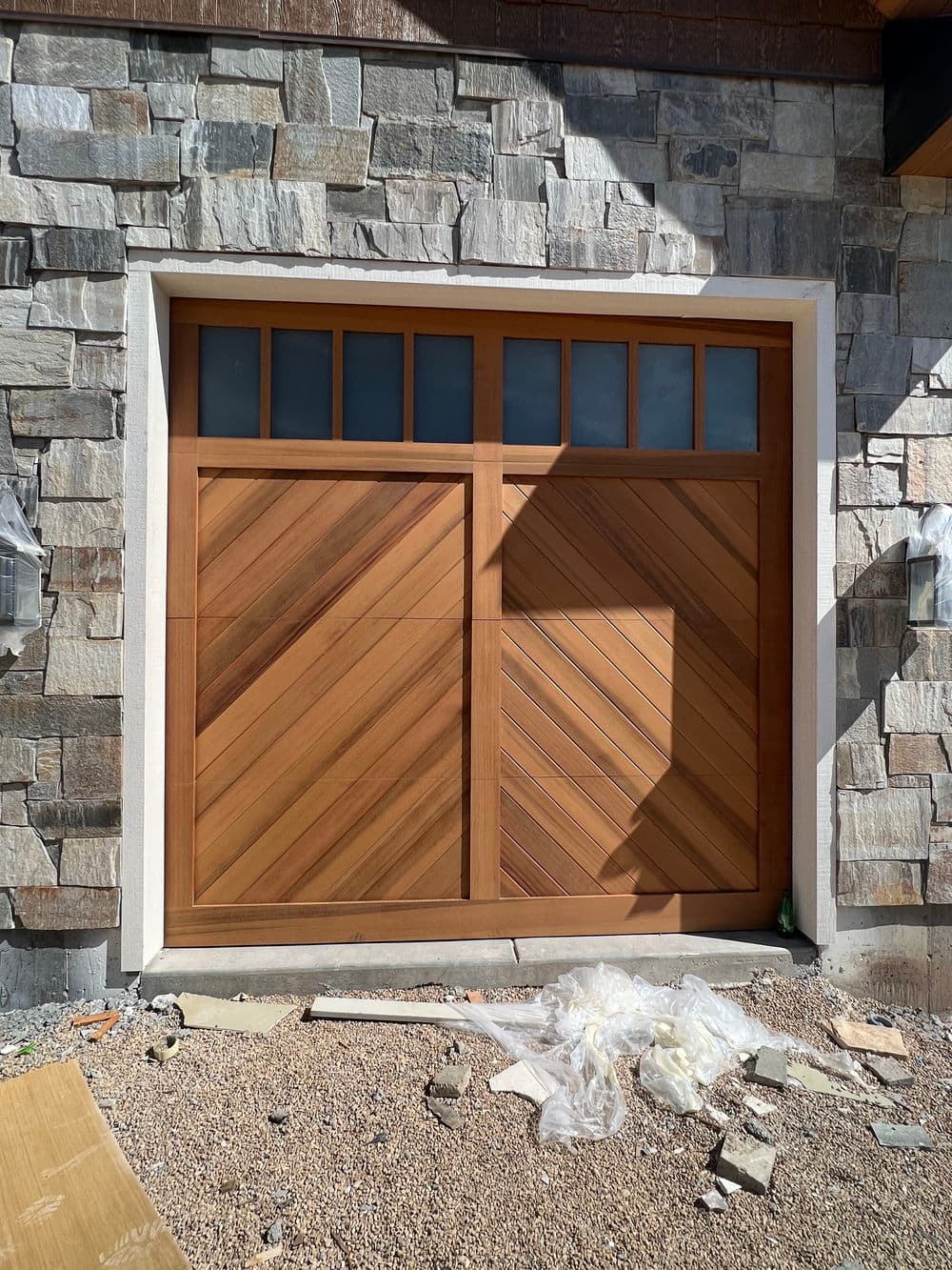 Modern wooden garage door with diagonal panels, framed by stone wall and construction debris.