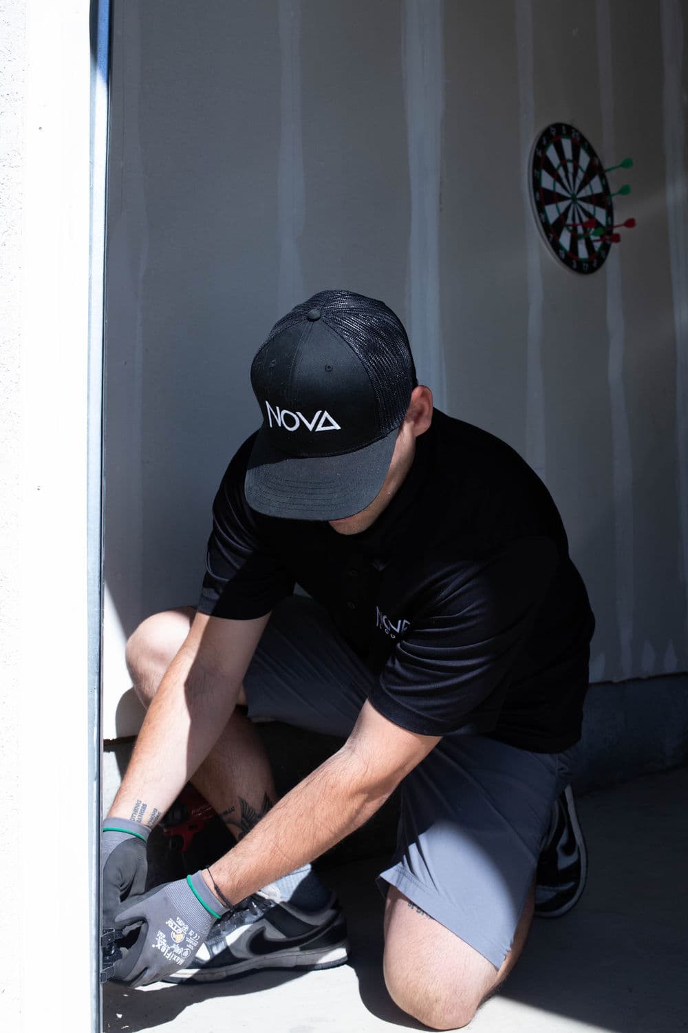 Technician wearing a NOVA cap repairs a door in a garage with a dartboard in the background.