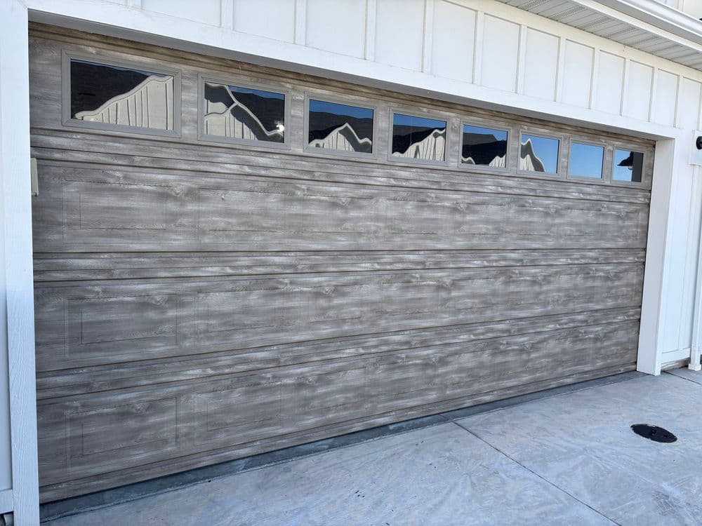 Modern grey wooden garage door with windows and horizontal paneling on a house exterior.