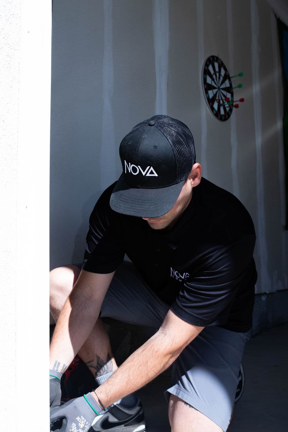 Worker in NOVA cap and shirt performing tasks in a garage with a dartboard in the background.